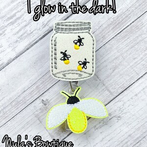 May include: A white mason jar with a gray lid filled with glowing yellow fireflies. The jar is attached to a clear plastic badge holder with a yellow firefly hanging below. The text "I glow in the dark!" is above the jar. The text "Myla's Boutique" is below the jar.