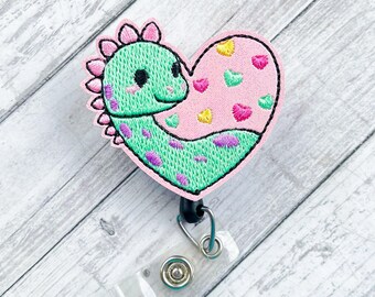 May include: A green and pink heart-shaped embroidered badge with a dinosaur design. The badge is attached to a clear plastic retractable badge holder with a black clip.