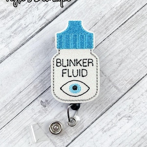 May include: A white and blue badge reel with the text "BLINKER FLUID" and an eye graphic. The badge reel has a blue top, a white body, and a black and silver retractable clip. The background is a light wood grain.