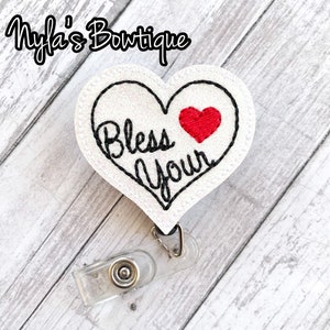 May include: A white heart-shaped badge reel with black stitched outline and the words "Bless Your" in black script. A small red heart is stitched on the right side. The badge reel has a clear plastic clip and retractable cord.