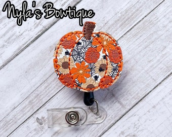May include: A white and orange pumpkin-shaped retractable badge holder with a floral and spiderweb pattern. The holder is attached to a clear plastic badge clip.