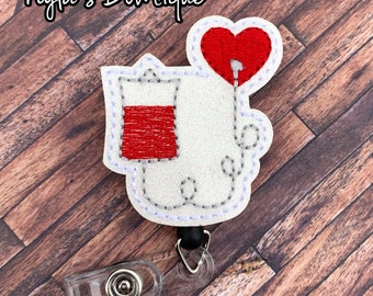 May include: A white and red badge reel featuring a heart and a blood bag design. The badge reel has a clear plastic clip and a black retractable cord. The text "Nyla's Boutique" is at the top of the image.