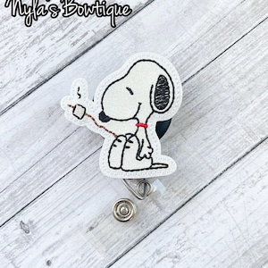 May include: A white Snoopy badge reel with black outline and a red collar, holding a marshmallow on a stick. The badge reel has a clear plastic retractable cord and a metal clip. The text "Nyla's Boutique" is at the top.