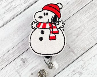 Könnte beinhalten: Ein Snoopy-Schneemann-Ausweis-Rollhalter mit roter Mütze und Schal. Der Ausweis-Rollhalter hat einen weißen Schneemannkörper mit schwarzen Knöpfen. Der Text "Nyla's Boutique" steht oben. Die Rolle hat ein einziehbares Kabel und einen Metallclip.