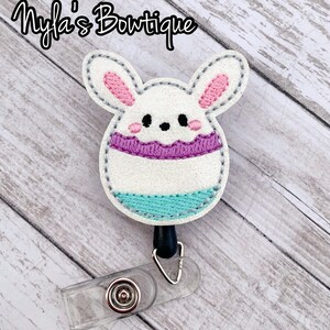 May include: White glitter Easter bunny egg with purple and blue accents, attached to a clear badge reel with a black retractable cord.  The bunny has pink ears and a cute face.  The text "Myla's Bowtique" is in the top left corner of the image.