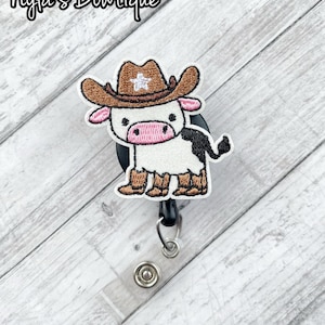 May include: A white cow with brown spots wearing a brown cowboy hat with a white star on it. The cow is wearing brown boots and has a black tail. The cow is attached to a black retractable badge reel.