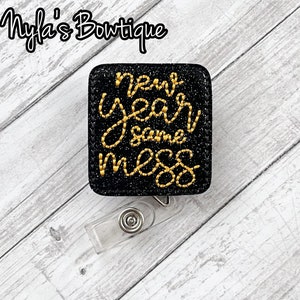 May include: A black, square badge reel with gold embroidered text that reads "new year same mess." The badge reel has a clear plastic clip and a silver metal button. The background is a light gray and white wood grain.