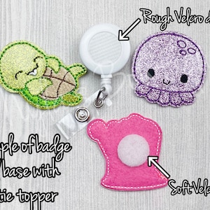 May include: Example of a badge reel base with a felt turtle topper. The reel has a white plastic base with a rough velcro dot. The turtle is green and yellow with a white face and black eyes. The felt topper is attached to the reel with a soft velcro dot.