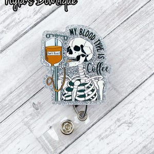 May include: A silver glitter badge reel with a skeleton drinking coffee from an IV bag. The IV bag says "Dark Roast". The text on the badge reel says "My blood type is coffee".
