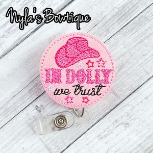 May include: A pink badge reel with the text "IN DOLLY we trust" and a pink cowboy hat design. The badge reel has a clear plastic clip and a retractable cord. The text "Nyla's Boutique" is at the top.