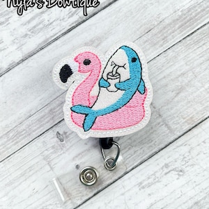 May include: A pink flamingo and blue shark embroidered badge reel. The shark is holding a drink. The badge reel has a clear plastic holder, a metal clip, and a snap closure. The text "Nyla's Boutique" is at the top.
