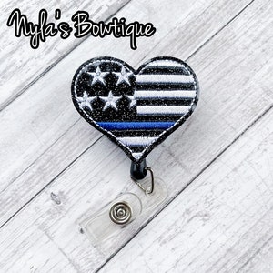 May include: A black and white heart-shaped badge holder with a thin blue line and an American flag design. The badge holder is attached to a clear plastic retractable badge reel.