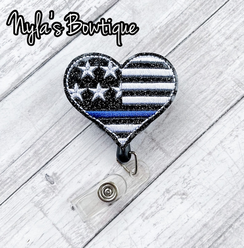 May include: A black and white heart-shaped badge holder with a thin blue line and an American flag design. The badge holder is attached to a clear plastic retractable badge reel.