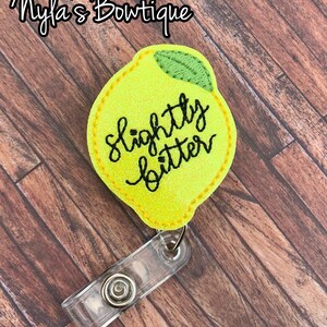 May include: A yellow lemon-shaped badge holder with the text "slightly bitter" embroidered in black. The badge holder has a clear plastic clip.