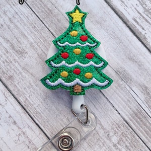 May include: A Christmas tree-shaped badge reel with a green glitter design, red and yellow ornaments, and a yellow star on top. The reel has a clear plastic strap and a metal clip. The text "Nyla's Bowtique" is in the upper left corner.