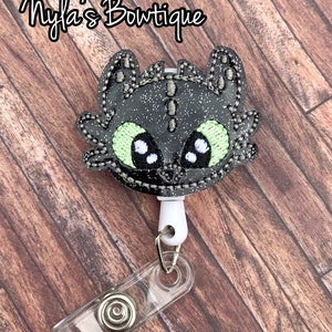 May include: Black and green glitter dragon character badge reel with a clear plastic clip.  "Myla's Boutique" is written in the top left corner of the image.