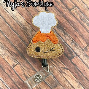 May include: A badge reel featuring a winking volcano design with a chef's hat. The volcano is orange and gold glitter, with a white hat. The text "Nyla's Boutique" is at the top. The reel has a clear plastic clip.