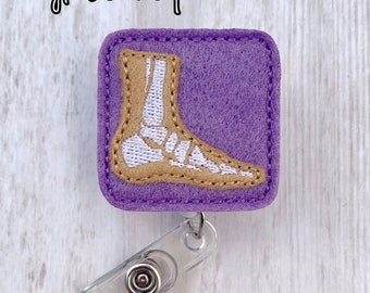 May include: Purple felt badge reel with a white embroidered image of a foot skeleton on a tan background. The text "Nyla's Boutique" is visible in the top left corner of the image.