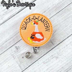 May include: A round orange badge with the text "QUACK-O-LANTERN" encircling a cartoon duck wearing a pumpkin costume. The badge has a yellow stitched border and a clear plastic retractable reel with a metal clip.