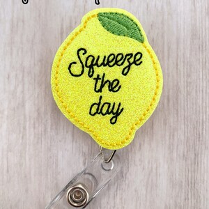 May include: Yellow lemon-shaped badge holder with the text "Squeeze the day" in black cursive lettering. The badge holder has a clear plastic clip.
