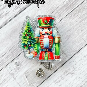 May include: A silver glitter badge reel with a Christmas nutcracker holding a green Christmas tree. The nutcracker is wearing a red and green uniform with a white beard and mustache.