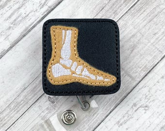 May include: Black and tan fabric badge holder with a white embroidered skeleton foot design.  The badge holder has a clear retractable badge reel.