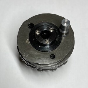 May include: A silver metal gear with a black center and a small silver pin on the side. The gear has a series of teeth around the edge.