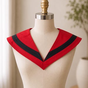 May include: A red collar with a black stripe detail, displayed on a mannequin. The collar has a V-neck design and is made of a smooth fabric. The mannequin has a gold-colored top and a neutral background.
