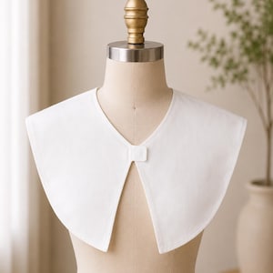 May include: A crisp white detachable collar featuring a wide, rounded Peter Pan style design. The collar is made of a smooth, solid white fabric and includes a small, rectangular tab closure at the center front neckline for a secure fit.