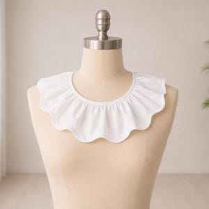 May include: A white, ruffled collar with a scalloped edge, displayed on a mannequin. The collar is made of a soft fabric and has a gathered design. The background includes a window and a potted plant.