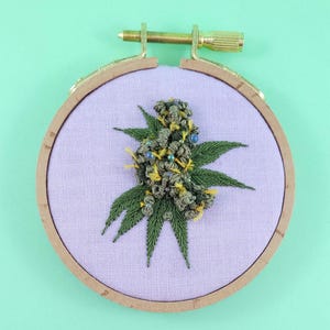 May include: An embroidered cannabis bud on a lavender fabric, set within a wooden embroidery hoop. The bud features green, yellow, and blue accents, with detailed green leaves. The background is a solid light green.