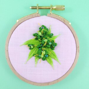 May include: An embroidered cannabis bud design on pink fabric, set within a wooden embroidery hoop. The bud is detailed with green thread and small blue beads. The hoop has a brass screw for tension adjustment. The hoop's diameter is approximately 10 cm.