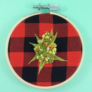 May include: Embroidered artwork in a wooden hoop. The fabric background has a red and black buffalo check pattern. The central design is a detailed embroidery of a green and yellow plant with white accents.