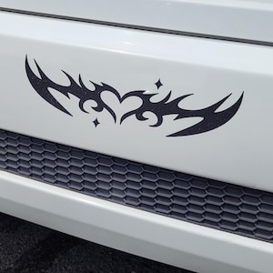 May include: A black tribal heart design car decal is affixed to a white vehicle bumper. The decal features a heart shape with stylized flame-like elements and star accents. The bumper has a black honeycomb grill.