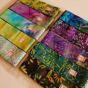 May include: Several folded fabric strips with vibrant patterns and numbered labels. The fabrics feature diverse designs, such as swirls, tie-dye, and floral motifs in shades of blue, green, purple, and yellow. Each strip is neatly folded.