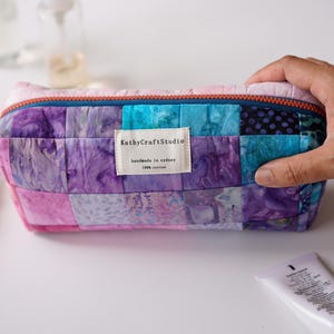 以下が含まれることがあります： ピンク、紫、ターコイズの色合いのパッチワークデザインの、長方形のキルティング化粧ポーチ。青いジッパーと「KathyCraftStudio」と書かれた小さな布タグが付いています。