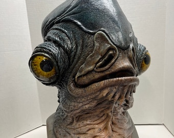 May include: A detailed, realistic mask of a creature with large, yellow eyes and a textured, gray-blue skin. The mask has a prominent brow, a wide mouth, and a detailed neck. The base of the mask is a gray color.