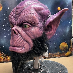 Könnte beinhalten: Eine lila und schwarze Alien-Kopfskulptur mit einem detaillierten Gesicht und großen Ohren. Die Skulptur steht auf einem schwarzen Sockel vor einem weißen Hintergrund.