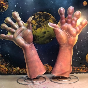 Könnte beinhalten: Zwei realistisch aussehende Prothesenhände mit jeweils fünf Fingern, montiert auf Metallständern. Die Hände sind fleischfarben und scheinen aus Silikon zu sein. Der Hintergrund ist dunkelblau mit gelben und orangefarbenen Planeten.