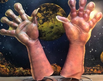 May include: Two realistic-looking prosthetic hands with five fingers each, mounted on metal stands. The hands are a fleshy pink color and appear to be made of silicone. The background is a dark blue with yellow and orange planets.