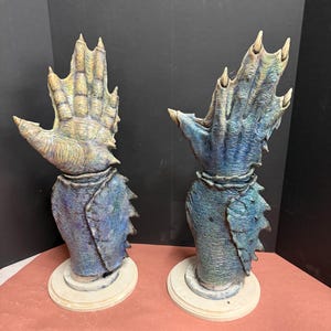 May include: Two sculptures of aquatic creature hands, each with a textured, multi-toned blue and tan finish. The hands are mounted on a blue base with a white, circular stand. The sculptures have a detailed, scaled appearance.