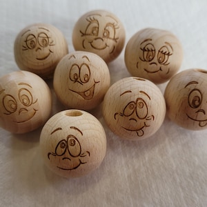 Könnte beinhalten: Acht Holzkugeln mit eingeätzten Cartoon-Gesichtern, die jeweils einen anderen Ausdruck zeigen. Die Gesichter sind in das hellbraune Holz geätzt, mit Merkmalen wie Lächeln und Zwinkern. Die Kugeln sind wahrscheinlich aus Naturholz.
