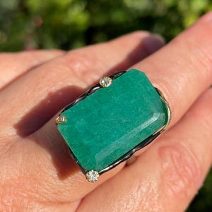 Könnte beinhalten: Ein großer Ring mit einem rechteckigen, smaragdgrünen Edelstein, Goldakzenten und zwei kleinen Diamanten. Der Ring hat einen schwarzen Rand und ist auf einem goldenen Band gefasst. Der Edelstein ist rechteckig und poliert.