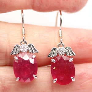 Puede incluir: Un par de pendientes colgantes de plata con piedras preciosas rojas oscuras de corte ovalado. Cada pendiente tiene un pequeño diseño de ángel con alas y un grupo de pequeñas piedras transparentes sobre la piedra roja. Los pendientes cuelgan de un gancho simple.