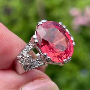 May include: A silver ring with a large, oval-shaped, bright pink gemstone. The ring's band has detailed embellishments with small, clear accent stones. The setting is ornate, with a scalloped edge around the main stone, creating a luxurious appearance.