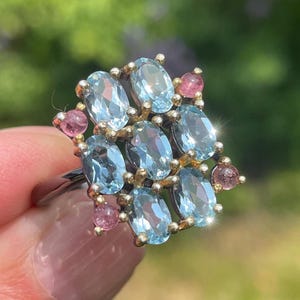 May include: A silver ring featuring a cluster of oval-cut, light blue gemstones surrounded by smaller pink gemstones. The ring has a gold-colored setting and is displayed against a blurred green background.