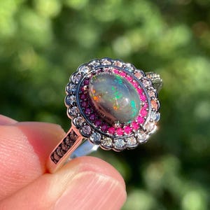 May include: A silver ring with an oval opal gemstone at its centre, displaying a play of colours. The opal is encircled by a halo of small pink gemstones and a border of clear stones. The ring's band has a decorative design.