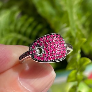 May include: A silver ring featuring a rectangular face encrusted with small, bright pink gemstones. The ring has a small, circular opening in the centre of the face. The band is silver with small black gemstones. The ring is set against green foliage.