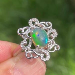 Puede incluir: Un anillo de plata con una piedra de ópalo ovalada con destellos iridiscentes. El ópalo está engastado en un diseño calado decorativo, adornado con pequeñas piedras de acento transparentes. El anillo se encuentra sobre un fondo verde borroso.