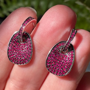 Könnte beinhalten: Ein Paar silberfarbene Ohrringe mit einer abgerundeten rechteckigen Form. Jeder Ohrring ist mit kleinen, leuchtend rosa Edelsteinen bedeckt. Die Ohrringe werden von einem kleinen, gebogenen Steg gehalten, der mit dem Hauptteil des Ohrrings verbunden ist.